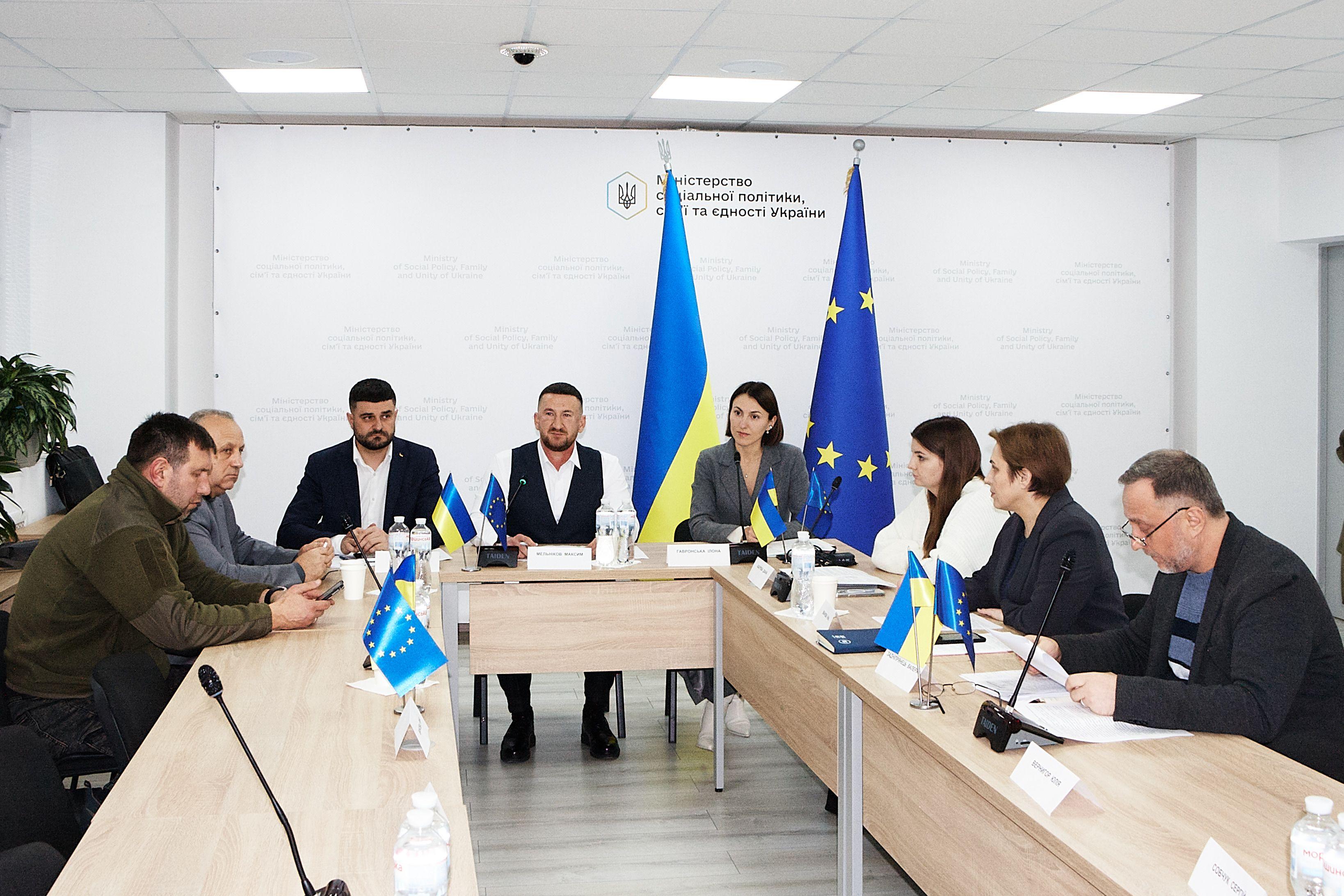 На фото чоловіки та жінки сидять за столом під час засідання Громадської ради На фото чоловіки та жінки сидять за столом під час засідання Громадської ради