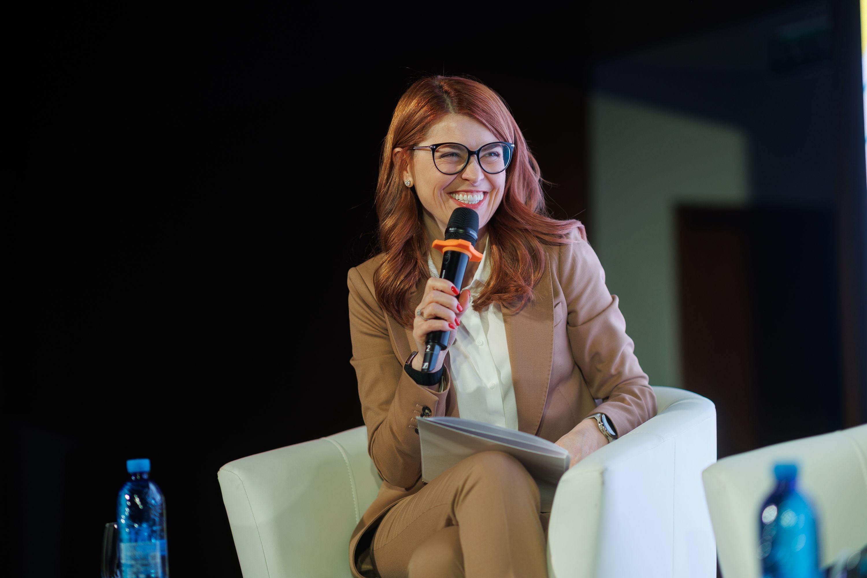 На фото жінка з мікрофоном виступає під час заходу На фото жінка з мікрофоном виступає під час заходу