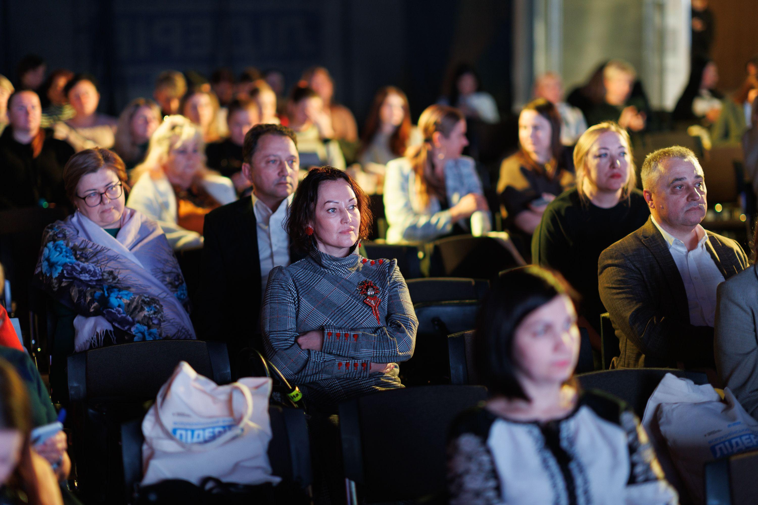 На фото жінки та чоловіки сидять в залі під час заходу На фото жінки та чоловіки сидять в залі під час заходу