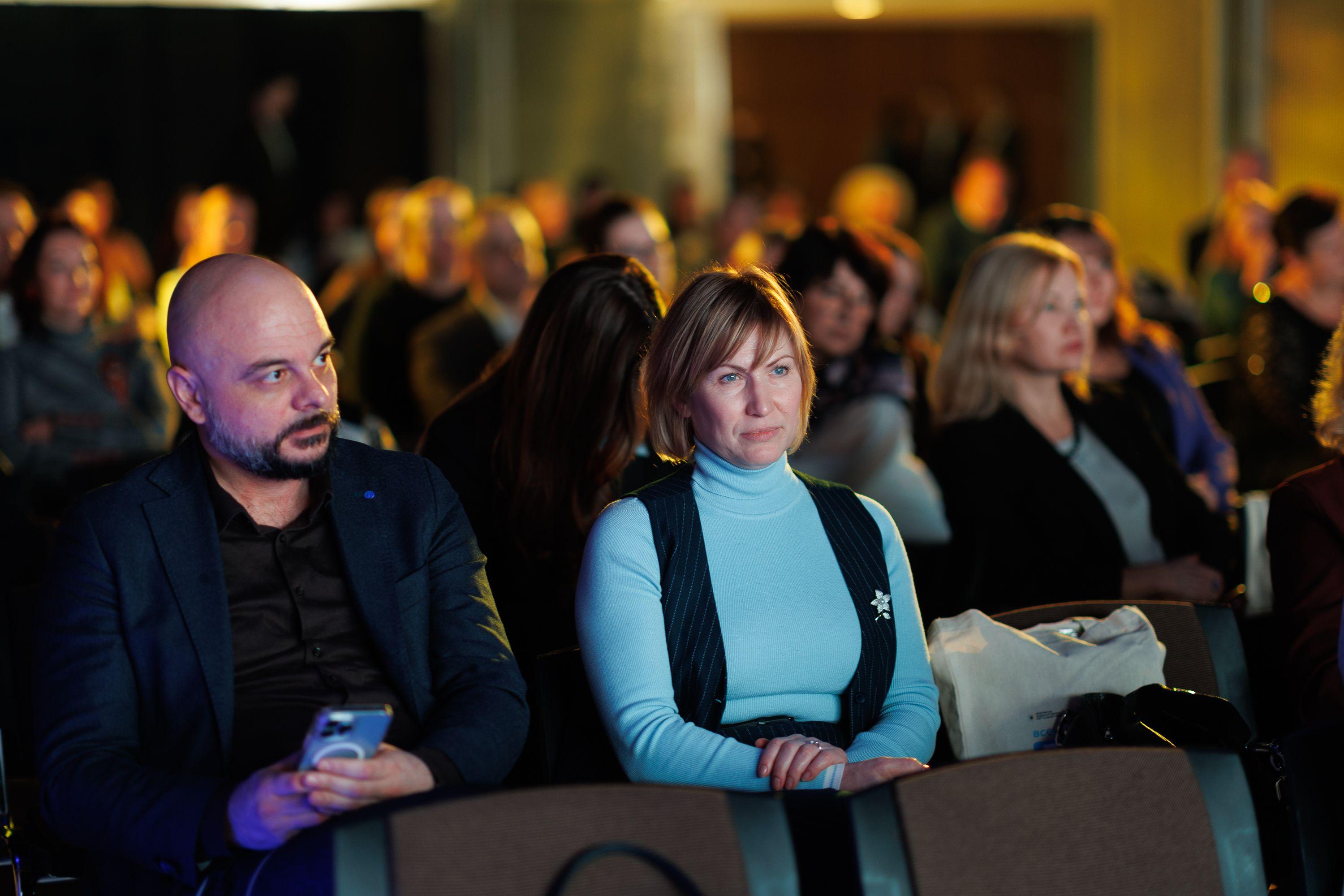 На фото жінки та чоловіки сидять в залі під час заходу На фото жінки та чоловіки сидять в залі під час заходу